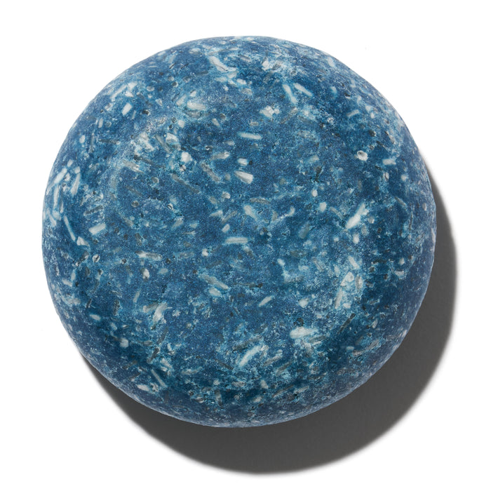 The Suds Shampoo Bar by NOT!CE Hair Co is a round, blue solid shampoo bar with white specks, set against a white background, representing sustainable self-care. It casts a soft shadow to the right, symbolizing an eco-friendly and plastic-free hair care option.