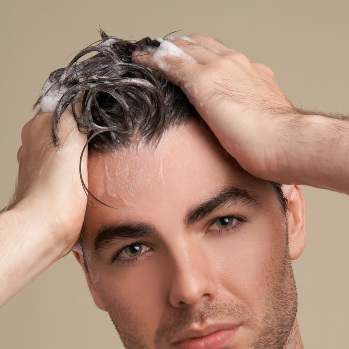 A person using Suds Shampoo Bar by NOT!CE Hair Co massages their scalp with lathered shampoo, hands visible, to promote hair growth. Their partially visible face against a neutral beige background captures the essence of sustainable self-care.