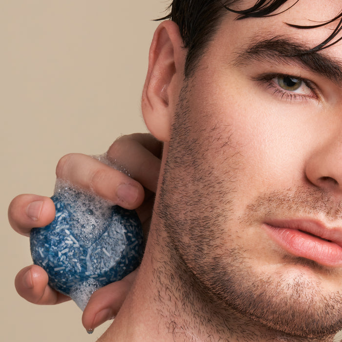 A person with wet, short hair and stubble holds the Suds Shampoo Bar by NOT!CE Hair Co to their cheek, producing a foamy lather. This plastic-free hair care scene highlights sustainable self-care against a neutral beige backdrop.