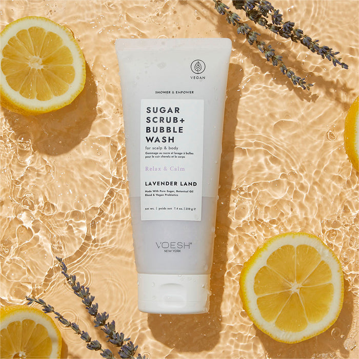 A tube of VOESH New Yorks Sugar Scrub + Bubble Wash - For Scalp and Body gently floats on rippling water, surrounded by lemon halves and lavender sprigs. The packaging highlights Relax & Calm with soothing vegan probiotics, emphasizing its natural tranquility.