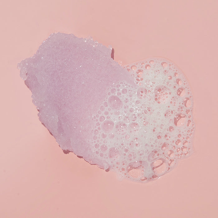 On a pink background, a bar of soap is covered in white bubbles, resembling VOESH New Yorks Sugar Scrub + Bubble Wash for Scalp and Body. Bubbles of varying sizes create a textured, frothy appearance around the soap.