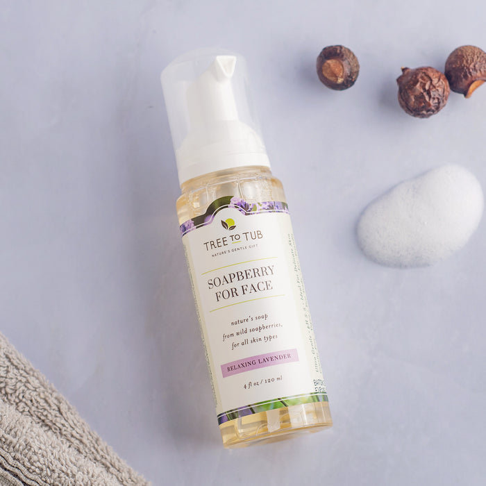 A bottle of Tree To Tubs Sulfate Free Hydrating Face Wash for Dry Skin rests on a light gray surface. Nearby, soapberries, soft white foam, and a folded gray towel create a clean, natural setting perfect for sensitive skin.