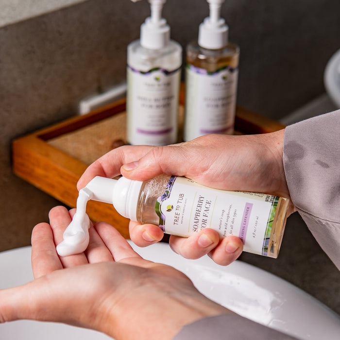 A person dispenses foaming soap for sensitive skin from a bottle labeled Tree To Tub into their hand over a bathroom sink. Two bottles of Sulfate Free Hydrating Face Wash for Dry Skin, featuring hydrating benefits, are visible in the background on a wooden tray.