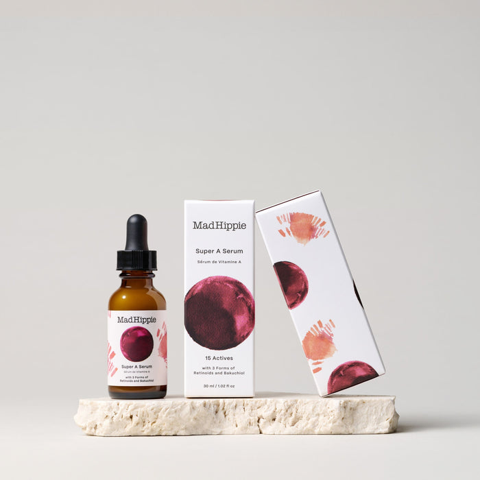 A Mad Hippie Super A Serum bottle and box sit on a stone surface. The packaging features red and pink abstract circles on white. An amber dropper bottle with a black cap aims to reduce wrinkles for youthful skin.