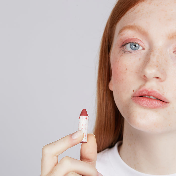 A person with long red hair and freckles holds a small tube from the Super Fan Pack by Axiology. They have light blue eyes and a white shirt, embodying vegan cosmetics elegance against a plain light gray background.
