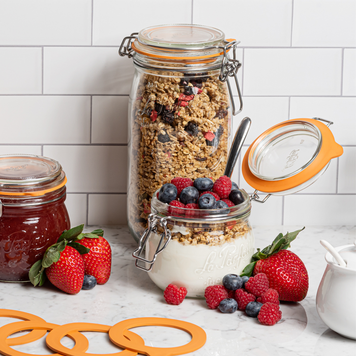 A countertop displays a Le Parfait Super Jar Variety Pack with one jar filled with granola and a smaller one layered with yogurt and berries. Nearby are strawberries, raspberries, blueberries, preserves in airtight-sealed jars, and orange sealing rings against a tiled backsplash.