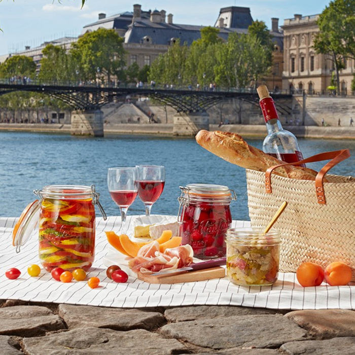 At a riverside picnic, a basket holds a baguette, wine with two glasses, and the Le Parfait Super Jar Variety Pack filled with pickled veggies. Melon slices and prosciutto are on a wooden board, with historic buildings and a bridge in the backdrop.
