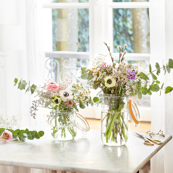 Two Le Parfait Super Jar Variety Pack canning jars, filled with assorted wildflowers such as eucalyptus, sit on a marble table. Scissors and loose leaves are nearby, with a large window in the background letting in natural light and offering a view of trees outside.