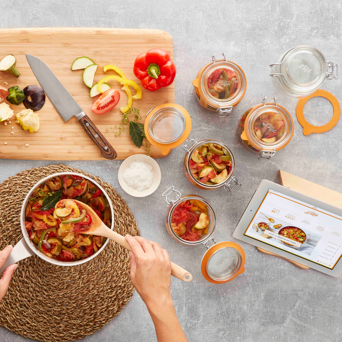 For vegetable ratatouille, a pot overflows with sliced veggies while more await their turn on a cutting board with a knife. Le Parfaits Super Jar Variety Pack, known for airtight seals, and an open tablet displaying the recipe complete the table setup.