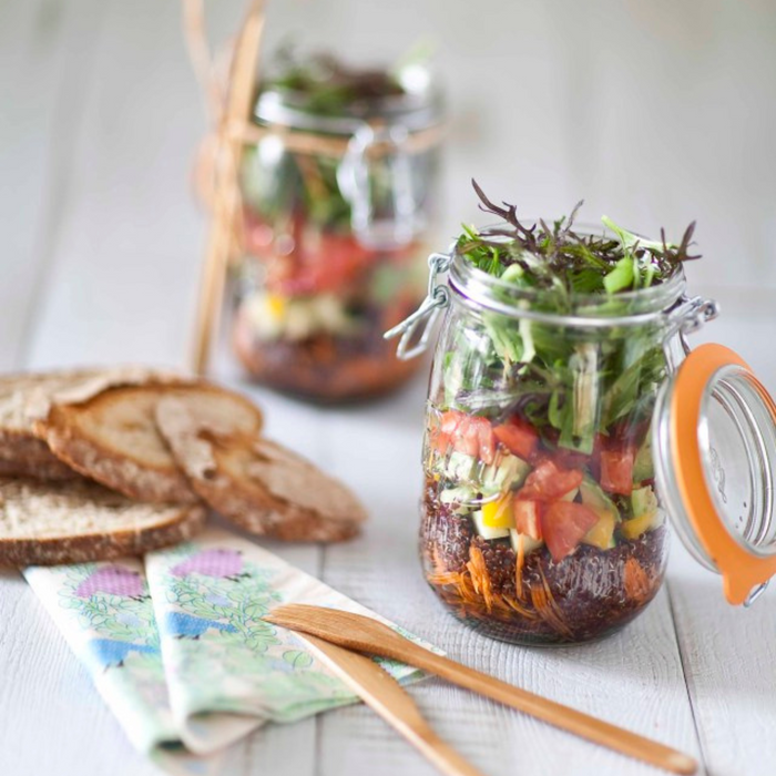 Two Le Parfait Super Jar Variety Pack jars filled with colorful layered salads, including greens, tomatoes, and assorted veggies, sit beside rustic bread slices and wooden utensils. The sealed canning jars are topped with herb sprigs on a light wooden table.