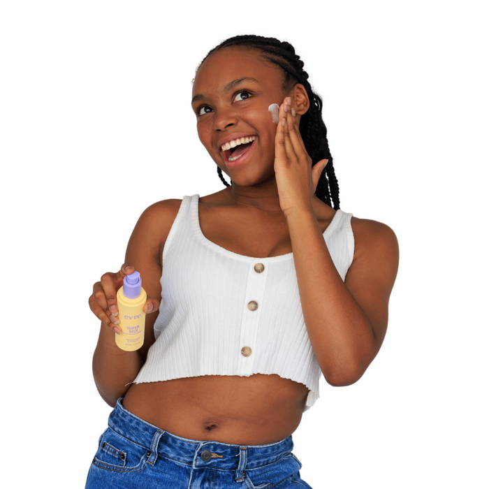 A smiling person with braided hair applies evre.s Super Nice Mānuka Honey & Vitamin E Face Moisturizer to their cheek. Clad in a white sleeveless top and blue jeans, they hold a pump dispenser bottle against a white background.