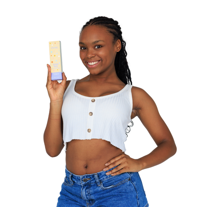 A person in a white crop top and blue jeans smiles while holding a yellow and purple box of Super Nice Mānuka Honey & Vitamin E Face Moisturizer by evre., against a plain white background.