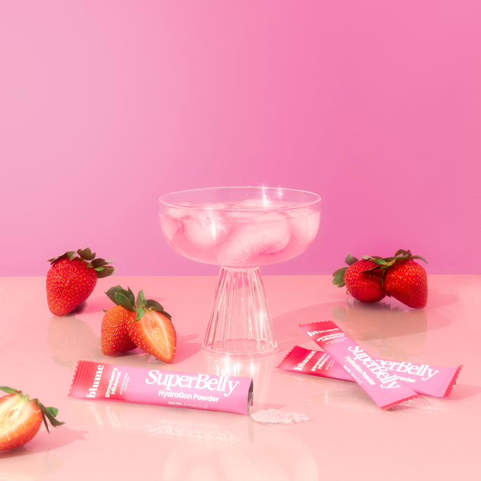 A glass of pink drink sits on a glossy surface, surrounded by strawberries and packets labeled SuperBelly Probiotic Hydration Powder by blume. The vibrant pink background and surface create a monochromatic theme celebrating gut health.