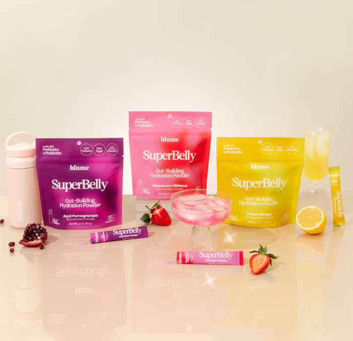 The display for Blumes SuperBelly Probiotic Hydration Powder features packaging in Acai Pomegranate, Strawberry Hibiscus, and Lemon Ginger. It includes a shaker bottle, a glass drink, and fruits like pomegranate, strawberries, and lemon—all promoting superfood-powered gut health.