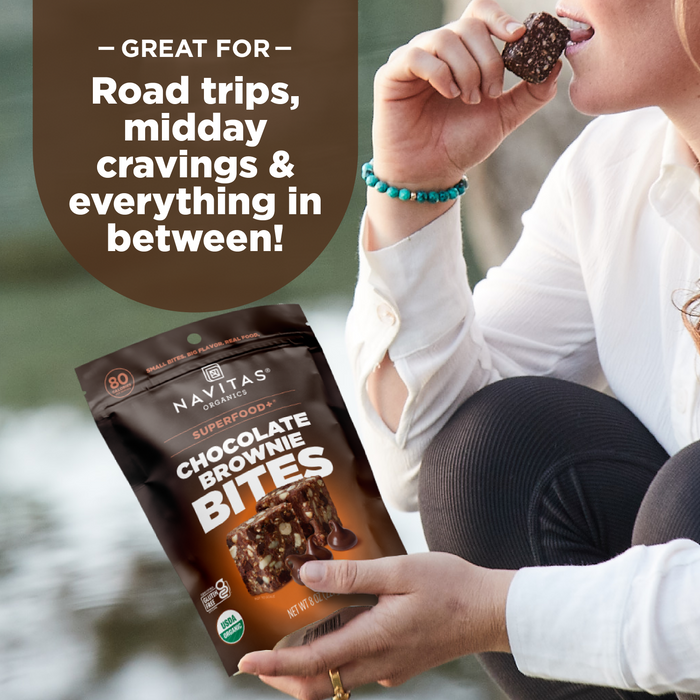 A person holds a Navitas Organics Superfood + Chocolate Brownie BITES pouch and enjoys a bite outdoors. Text above says: Perfect for road trips, midday cravings & more! Made with rich dark chocolate and organic cacao.
