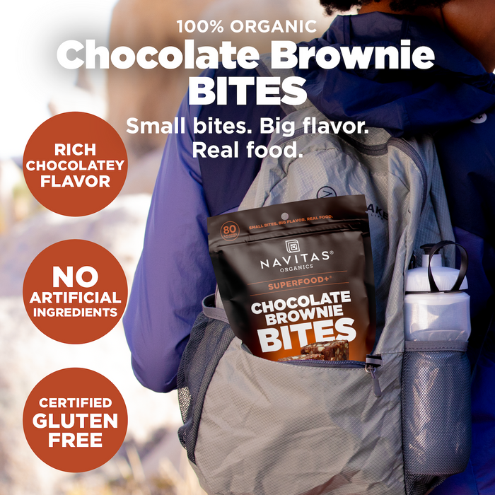 A person outdoors with a backpack pocket holding Navitas Organics Superfood + Chocolate Brownie BITES. Text highlights organic cacao's rich chocolatey flavor, gluten-free, no artificial ingredients, and "Small bites. Big flavor. Real food.