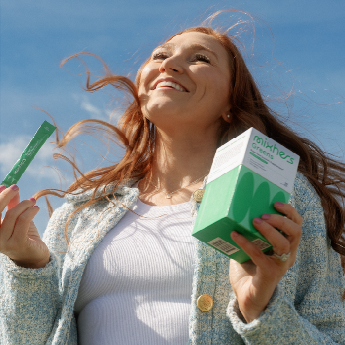 A woman with long red hair holds a box of Mixhers Superfood Greens Supplement Powder and a small packet. Smiling at the bright blue sky, shes in a light jacket and white top, her hair tousled by the wind, embodying natural detoxification and vibrant womens nutrition.