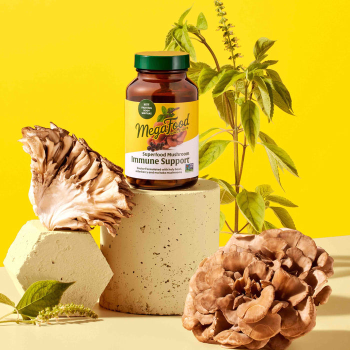 A jar of MegaFood Superfood Mushroom - Immune Support rests on a cylindrical pedestal with large mushrooms and green plants against a bright yellow background. It features Maitake mushroom and Holy Basil.