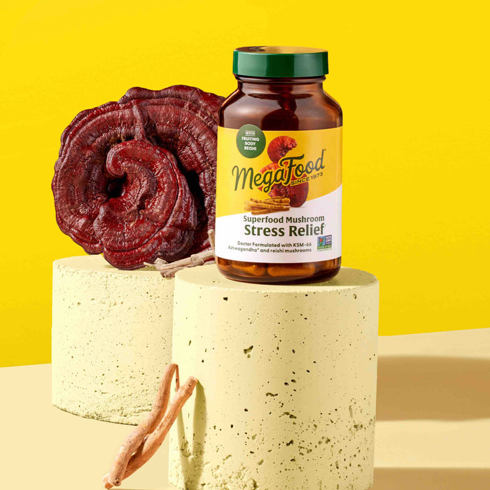 A bottle of MegaFoods Superfood Mushroom - Stress Relief, with soothing Reishi, is placed on a beige pedestal against a yellow background. Beside it are a large mushroom and two smaller slices, highlighting its adaptogenic benefits.