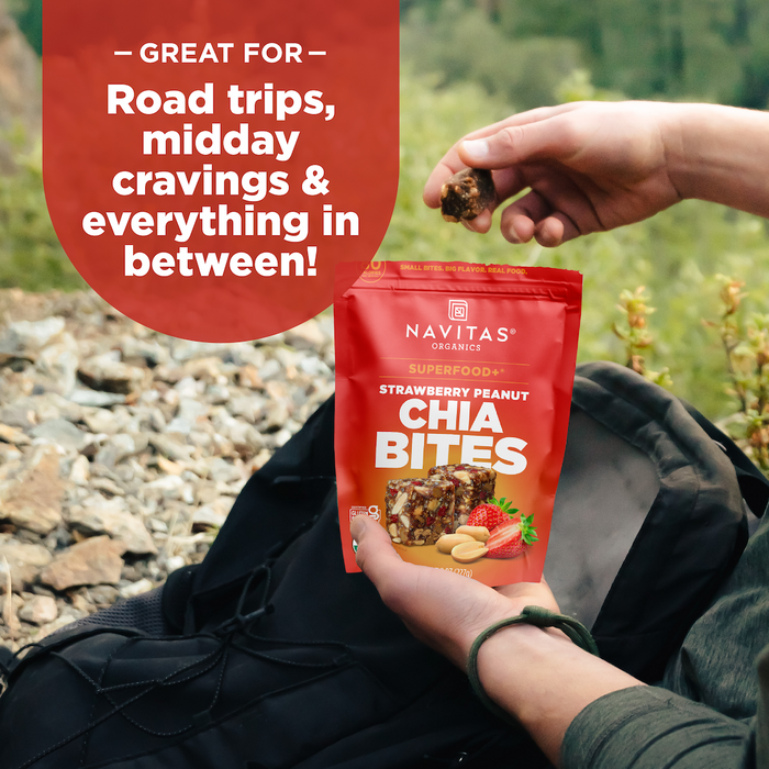 Outdoors, someone holds a Navitas Organics Superfood+ Strawberry Peanut Chia BITES bag and a snack. Text says: “Superfood snacks—perfect for road trips, midday cravings & more!”.