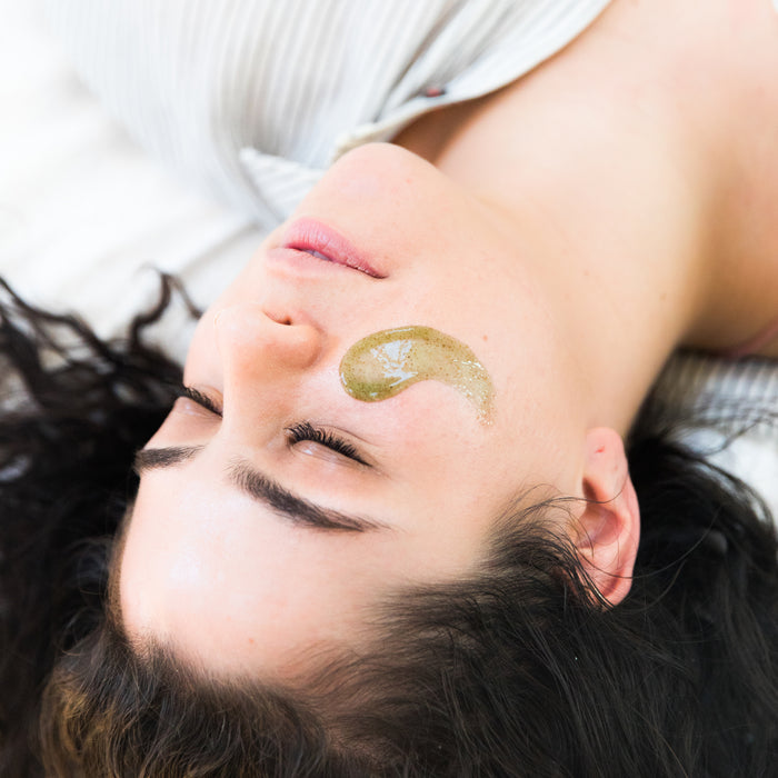 A person with long, dark hair lies on their back with a Superfruits & Calendula Facial Scrub by Beauty by Earth applied to their right cheek. They appear relaxed and wear a light-striped top.