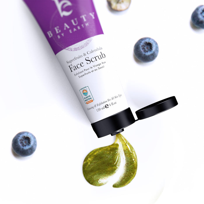 A tube of Beauty by Earths Superfruits & Calendula Facial Scrub spills green scrub onto a smooth surface, surrounded by scattered blueberries. The tube lies angled with its black cap open.