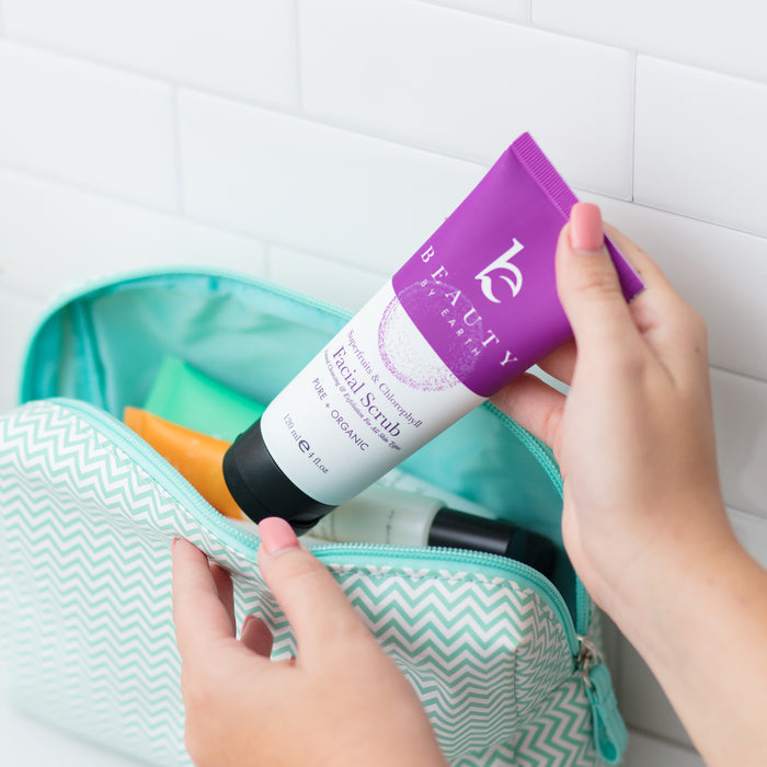 Someone holds a purple tube of Superfruits & Calendula Facial Scrub by Beauty by Earth over a light blue zigzag-patterned toiletry bag filled with other superfruit-infused products on a white tile background.