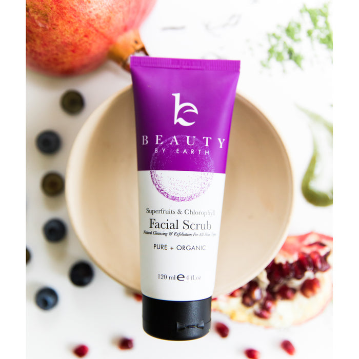 A 120 ml purple and white tube of Beauty by Earths Superfruits & Calendula Facial Scrub, known for its exfoliating power, sits on a bowl surrounded by vibrant pomegranate seeds, half a pomegranate, blueberries, and lush green leaves.