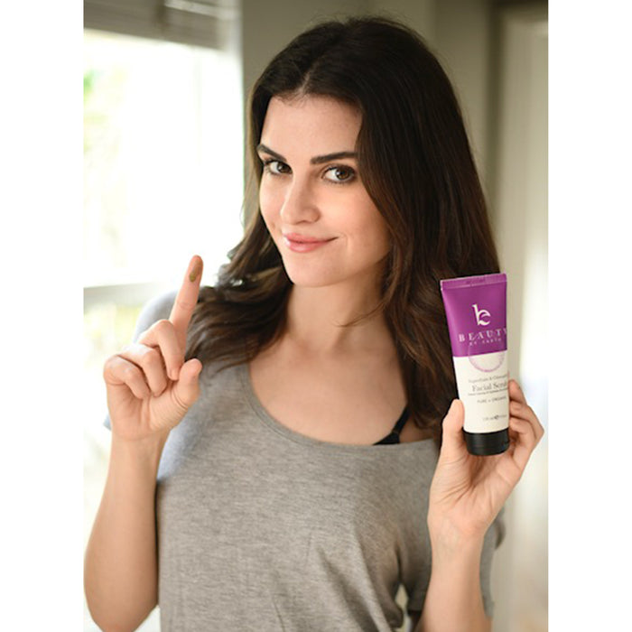 A woman with long, dark hair holds a tube of Beauty by Earths Superfruits & Calendula Facial Scrub, pointing to a dab on her finger. She wears a gray top and stands near a window.