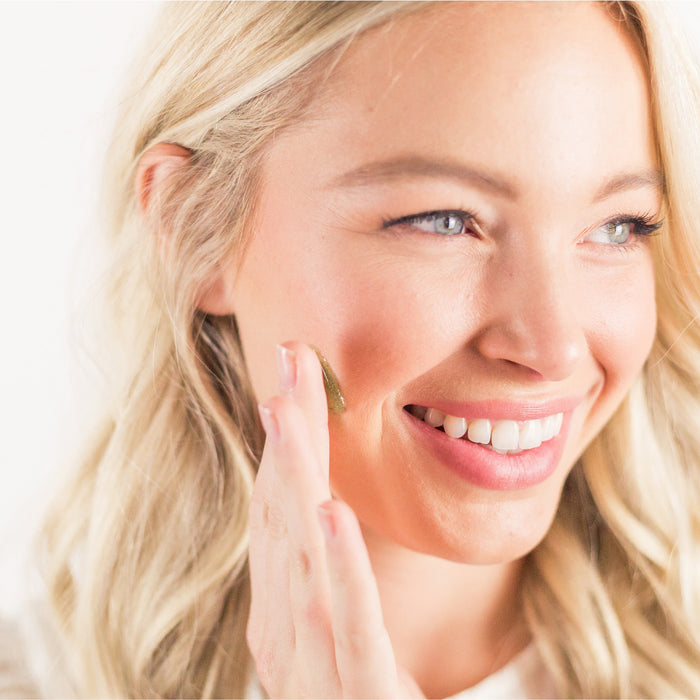 A person with long blonde hair smiles while applying Beauty by Earths Superfruits & Calendula Facial Scrub to their cheek with their fingertips. The softly blurred light background highlights their radiant face.