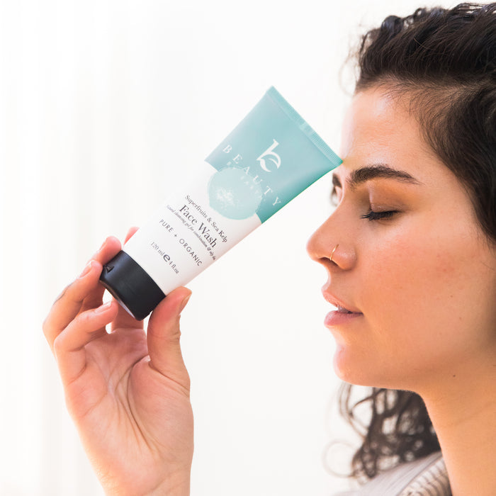 A person with curly hair holds a teal and white tube of Beauty by Earths Superfruits & Sea Kelp Facial Cleanser near their nose, eyes closed. The brightening and pore refining product stands out against the softly blurred background.