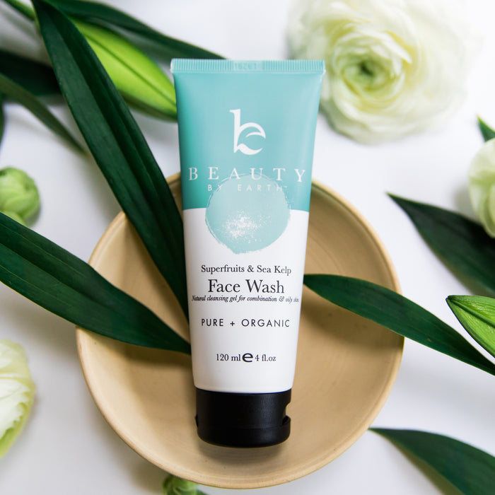 On a white surface, surrounded by green leaves and delicate white flowers, rests a tube of Beauty by Earths Superfruits & Sea Kelp Facial Cleanser. The label emphasizes its pure, organic formulation perfect for combination to oily skin.