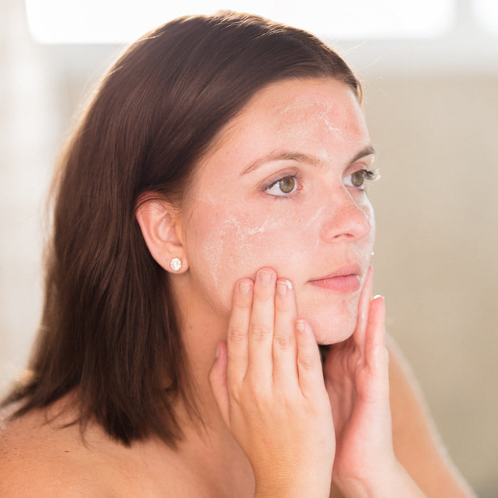 A woman with wet hair uses Beauty by Earths Superfruits & Sea Kelp Facial Cleanser, applying the foamy wash to her face with both hands. She gazes upwards against a blurred background, evoking a bathroom setting.