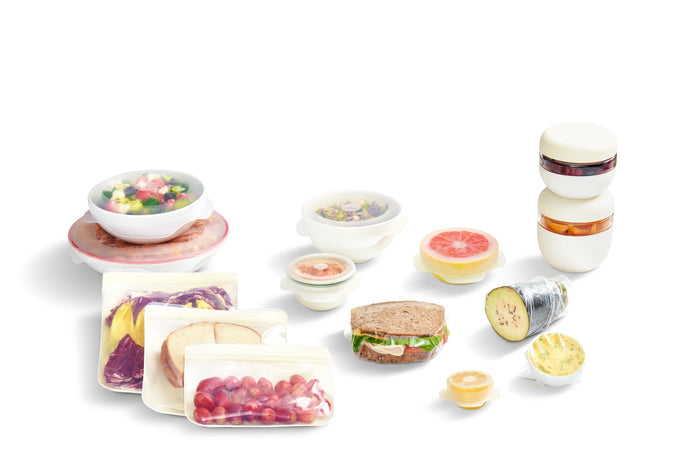 The W&P Sustainable Kitchen Set includes a colorful display of packed lunches with reusable stretch lids, featuring salads, sandwiches, snacks, and fruit in clear containers. Slices of bread and grapes are artfully arranged on a white background for an eco-friendly solution.