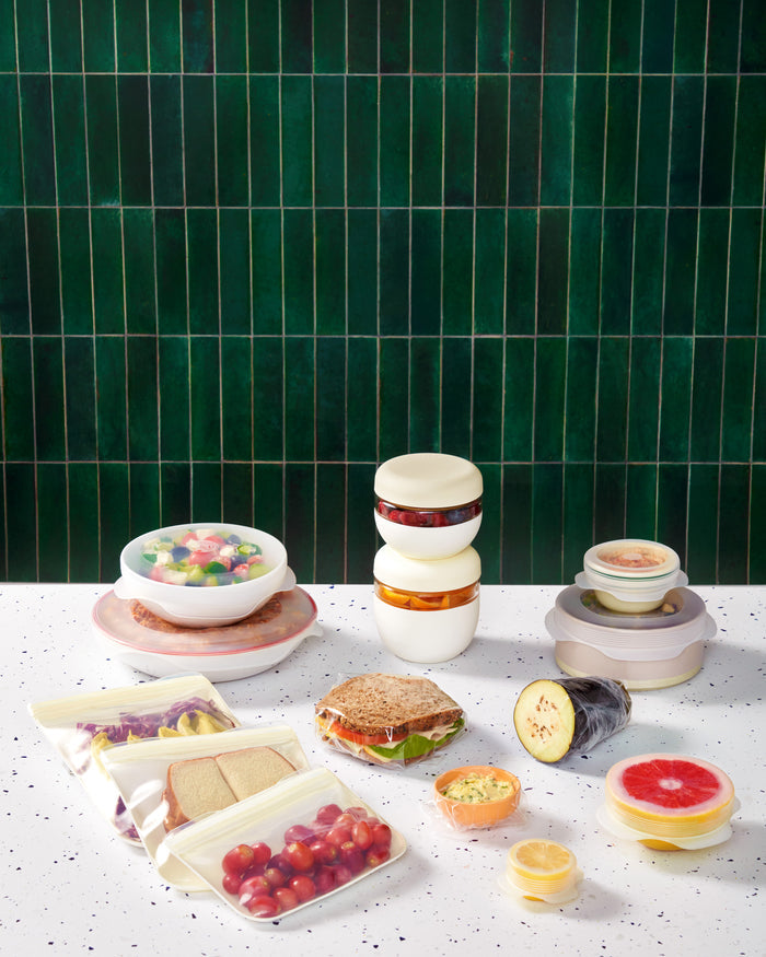 On a speckled countertop, W&Ps Sustainable Kitchen Set features stacked borosilicate glass containers with reusable stretch lids, holding fruits, a sandwich, vegetables, and sauces. A dark green tiled wall in the background completes this eco-friendly kitchen scene.