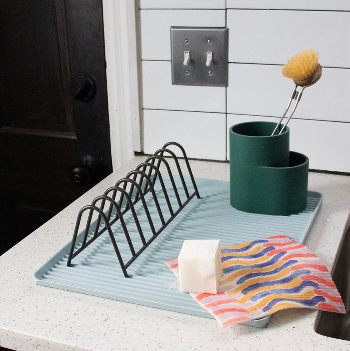 A kitchen countertop displays eco cleaning essentials: a drying rack on a ribbed mat, a sponge, and scrub brush. Two green containers hold a cleaning brush, while Supra Endura Swedish Dishcloths sit beside a super absorbent white sponge. A striped cloth is neatly spread out nearby.