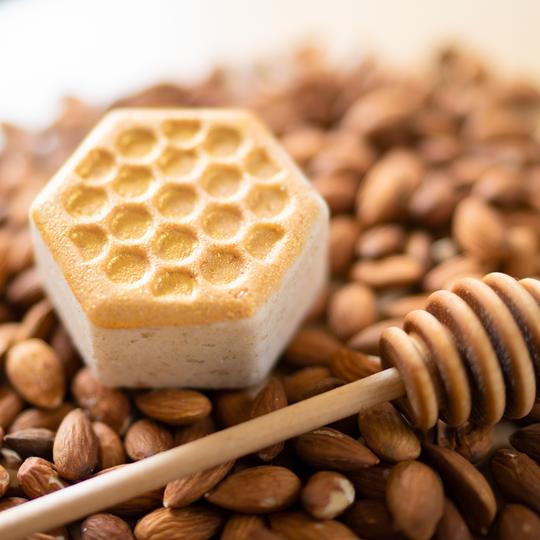 The Sweet Honey Almond Froth Bomb by Pacha Soap Co., hexagonal with a honeycomb pattern, rests on almonds. A wooden honey dipper hints at beekeeper-direct honey in the softly lit scene.