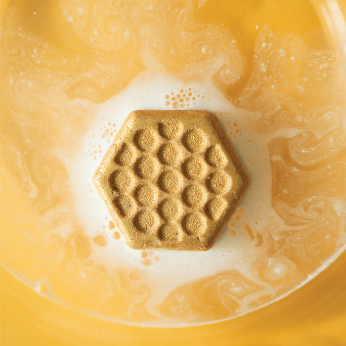 The Sweet Honey Almond Froth Bomb by Pacha Soap Co. is a golden, hexagonal bath bomb with a honeycomb pattern that creates swirling yellow and white water for a natural, cruelty-free and luxurious soak.