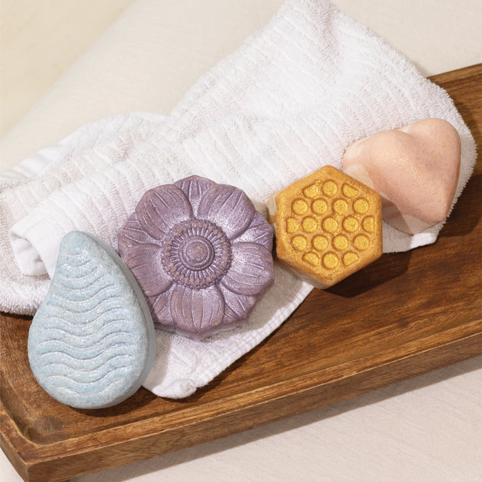 Four uniquely shaped and colored soaps—including a blue teardrop, a purple flower, a pink heart, and a yellow honeycomb inspired by Pacha Soap Co.'s Sweet Honey Almond Froth Bomb—rest on a white towel atop a wooden tray.