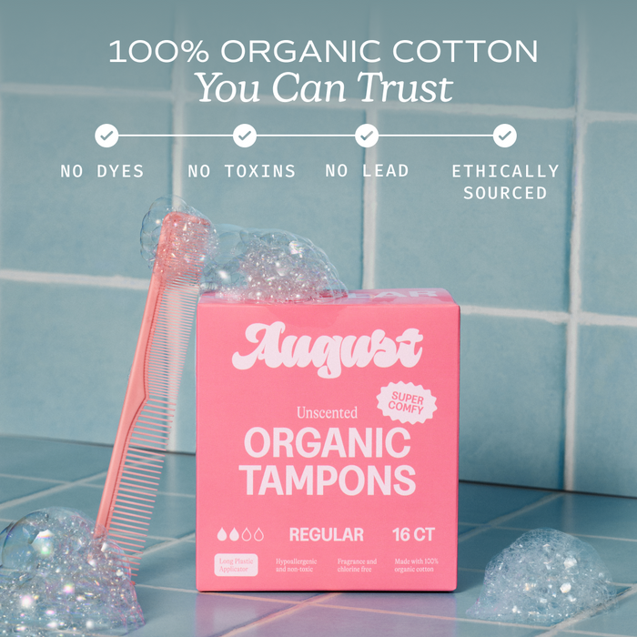 A pink box of August Regular Tampon (16 count) sits on blue tiles with a pink comb and bubbles. These unscented, hypoallergenic tampons include a BPA-free applicator and are free from dyes and toxins.