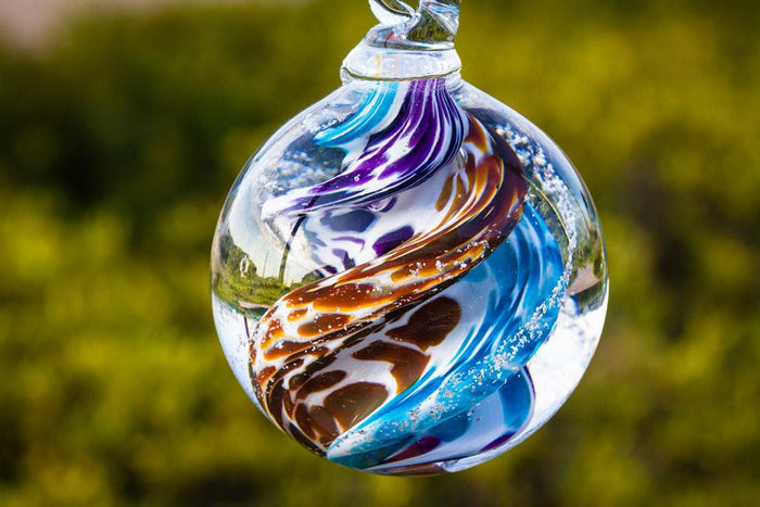 Tan and purple cat eye cremation orb with swirled colors hanging outdoors against a green backdrop