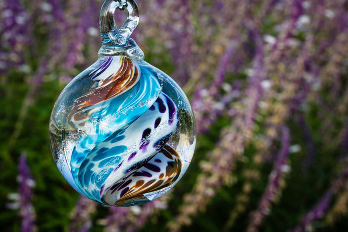 Multicolored glass cremation orb with swirling blue teal brown and purple patterns hanging from a loop in a garden setting.