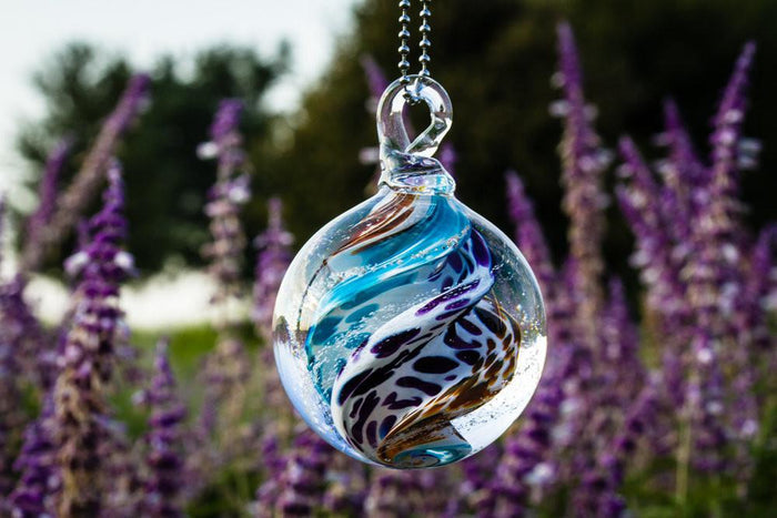 Close up of a multicolored glass cremation orb with blue purple brown white swirls hanging from a metal chain against a garden background.
