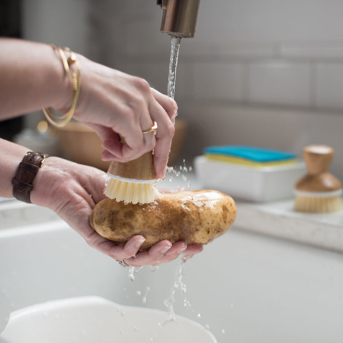 Using the Full Circle Tater Mate - Eye-Removing Potato Scrubber, a person thoroughly cleans a potato under running water at the kitchen sink. Hands grip it firmly for effective cleaning. In the background, a soap dispenser and cleaning brush signal the start of meal preparation.