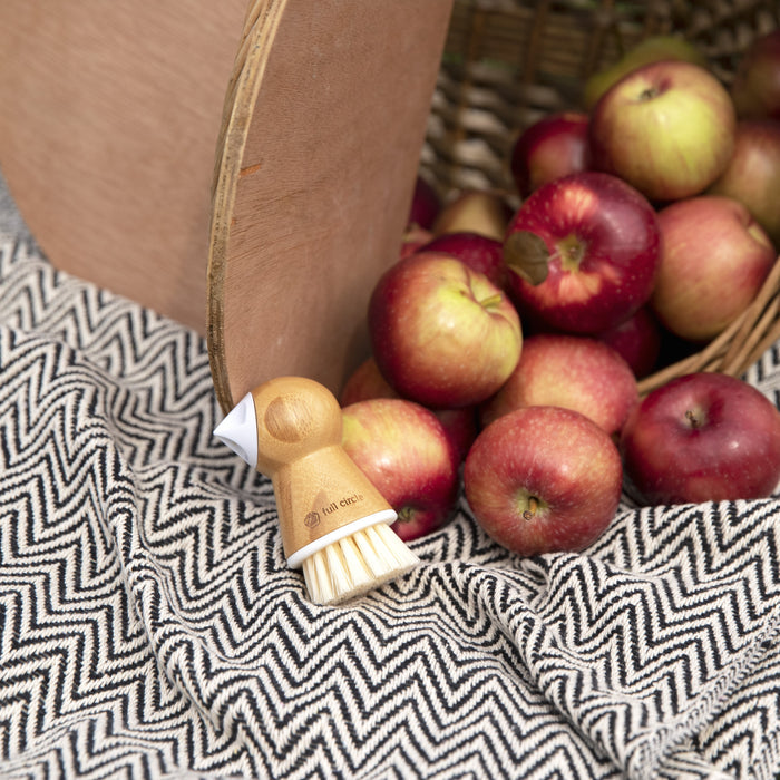 Red and green apples spill from a wicker basket onto a black-and-white chevron-patterned cloth, next to the Full Circle Tater Mate - Eye-Removing Potato Scrubber with bristles facing downward, ready for meal preparation.