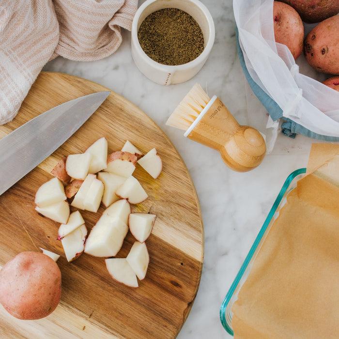 A wooden cutting board holds diced potatoes and a knife. Beside whole potatoes in a blue bag rests the Tater Mate Eye-Removing Potato Scrubber from Full Circle, near a jar of herbs and a parchment-lined baking dish, all on marble with a beige cloth.