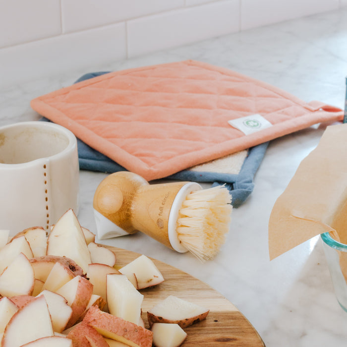 Chopped potatoes on a wooden board with Full Circles Tater Mate Eye-Removing Potato Scrubber nearby, an orange pot holder folded, and a ceramic jug, all atop a marble countertop.