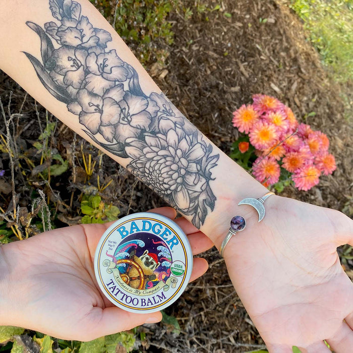 A person showcases a floral tattoo on their forearm, adorned with a silver bracelet featuring crescent charms, while holding a tin of Badger Balms Tattoo Balm next to pink flowers for stylish and organic aftercare.