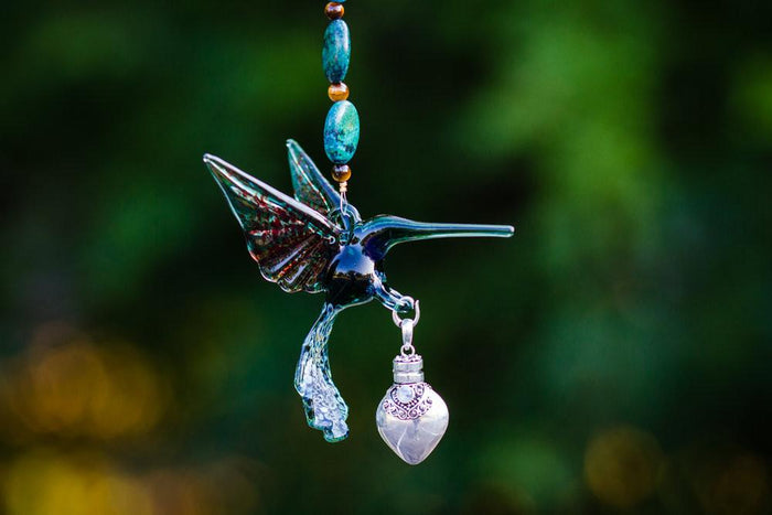 Close up of teal glass hummingbird with turquoise beads and a silver heart keepsake urn hanging below against a green blurred background.