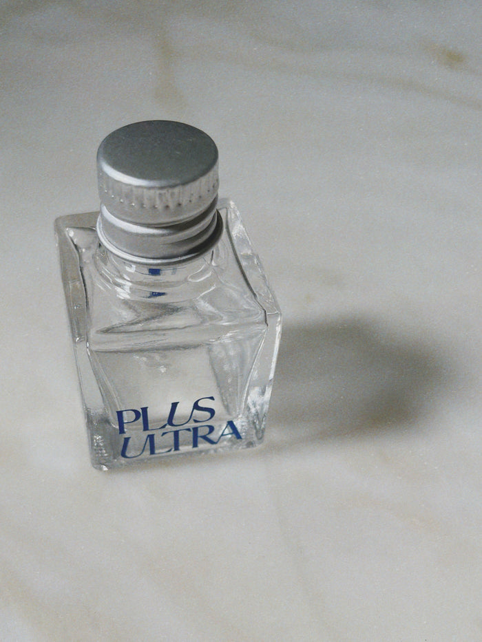 A small, square glass perfume bottle with a silver cap labeled Plus Ultra in blue lettering rests elegantly on a light marble surface, as meticulously designed as Plus Ultras Teeth Whitening Gel with Bamboo Applicator, casting a soft shadow.